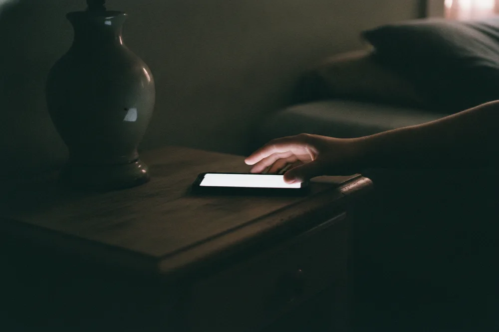 Hand reaching for a glowing phone on a nightstand in a dark bedroom - the after-hours call